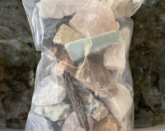 May include: A clear plastic bag filled with a variety of raw gemstones. The stones are in shades of pink, white, green, and brown. A long, dark, needle-like crystal is visible among the stones.