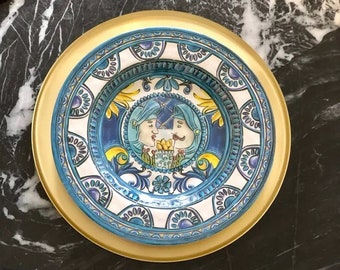 May include: A blue and white ceramic plate with a gold rim. The plate features a colorful design of two people with blue turbans and yellow accents. The plate is on a black marble surface.
