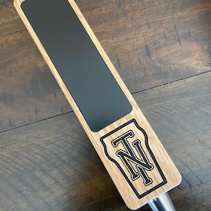Op de afbeelding: Een houten biertap met een zwart schoolbord oppervlak en een zwart-wit gegraveerd "TN" logo.