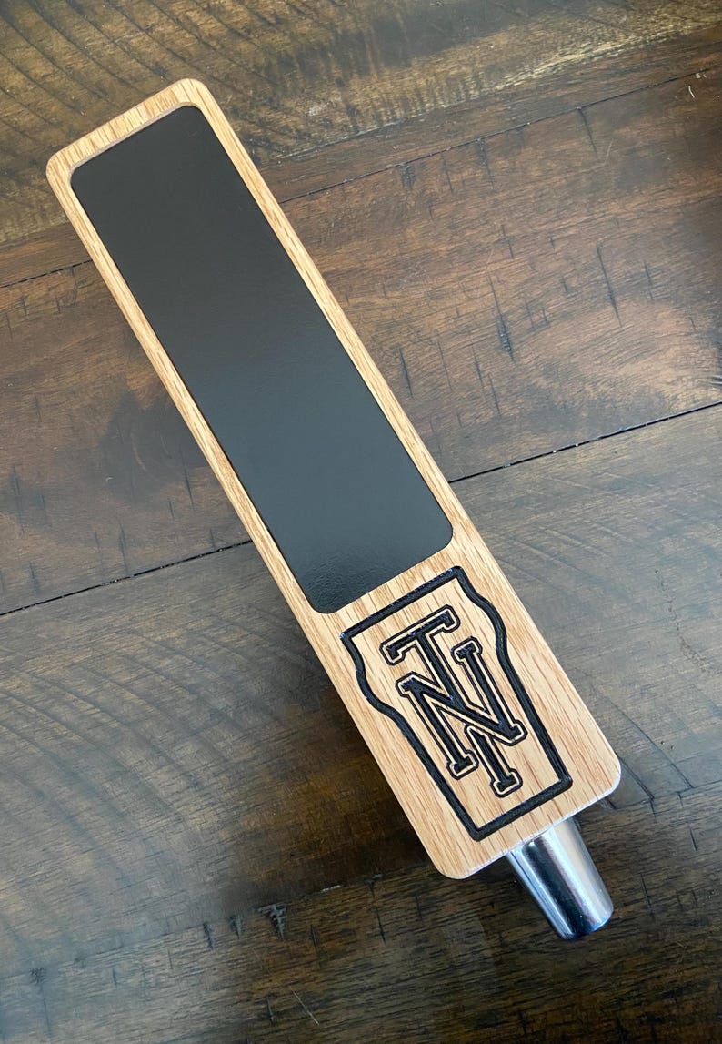 Op de afbeelding: Een houten biertap met een zwart schoolbord oppervlak en een zwart-wit gegraveerd "TN" logo.