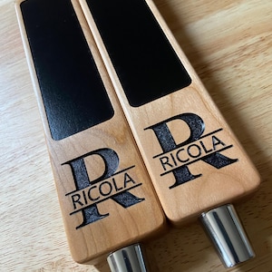 Op de afbeelding: Twee houten biertapkranen met zwarte schoolbord-inzetstukken. Elke kraan is gepersonaliseerd met de letter "R" en de naam "Riccola".