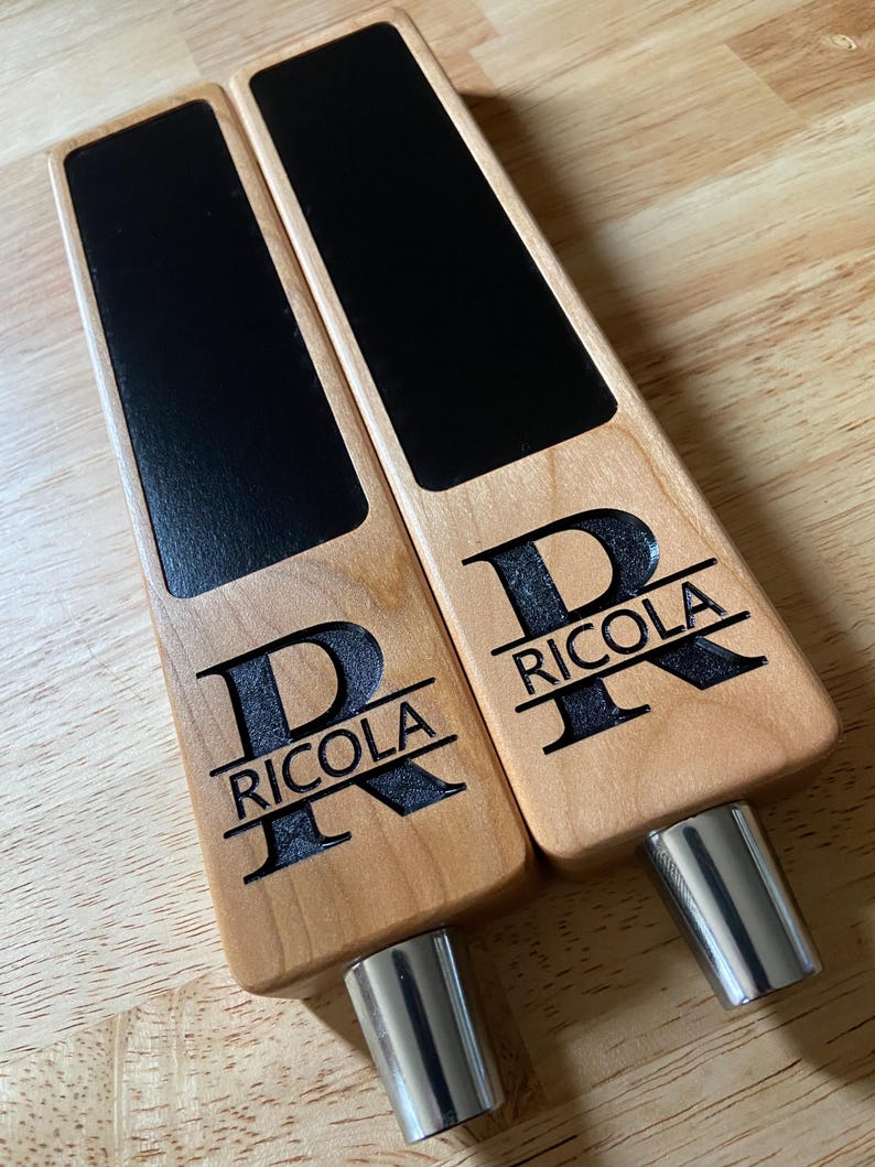 Op de afbeelding: Twee houten biertapkranen met zwarte schoolbord-inzetstukken. Elke kraan is gepersonaliseerd met de letter "R" en de naam "Riccola".