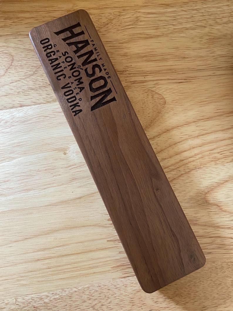 Op de afbeelding: Een houten bord met de tekst "HANSON of SONOMA GRAPE BASED ORGANIC VODKA FAMILY MADE" erop gegraveerd.