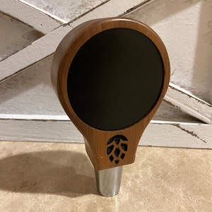 May include: A wooden beer tap handle with a black chalkboard insert and a carved hop design.