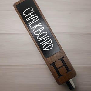 Op de afbeelding: Een houten biertap met een zwarte krijtbord-inzet met de tekst "CHALKBOARD" en een gegraveerde letter "H" aan de zijkant.