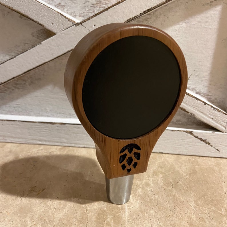 May include: A wooden beer tap handle with a black chalkboard insert and a carved hop design.