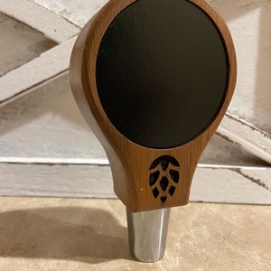 May include: A wooden beer tap handle with a black circular insert and a carved hop design. The handle is brown and has a silver metal base.