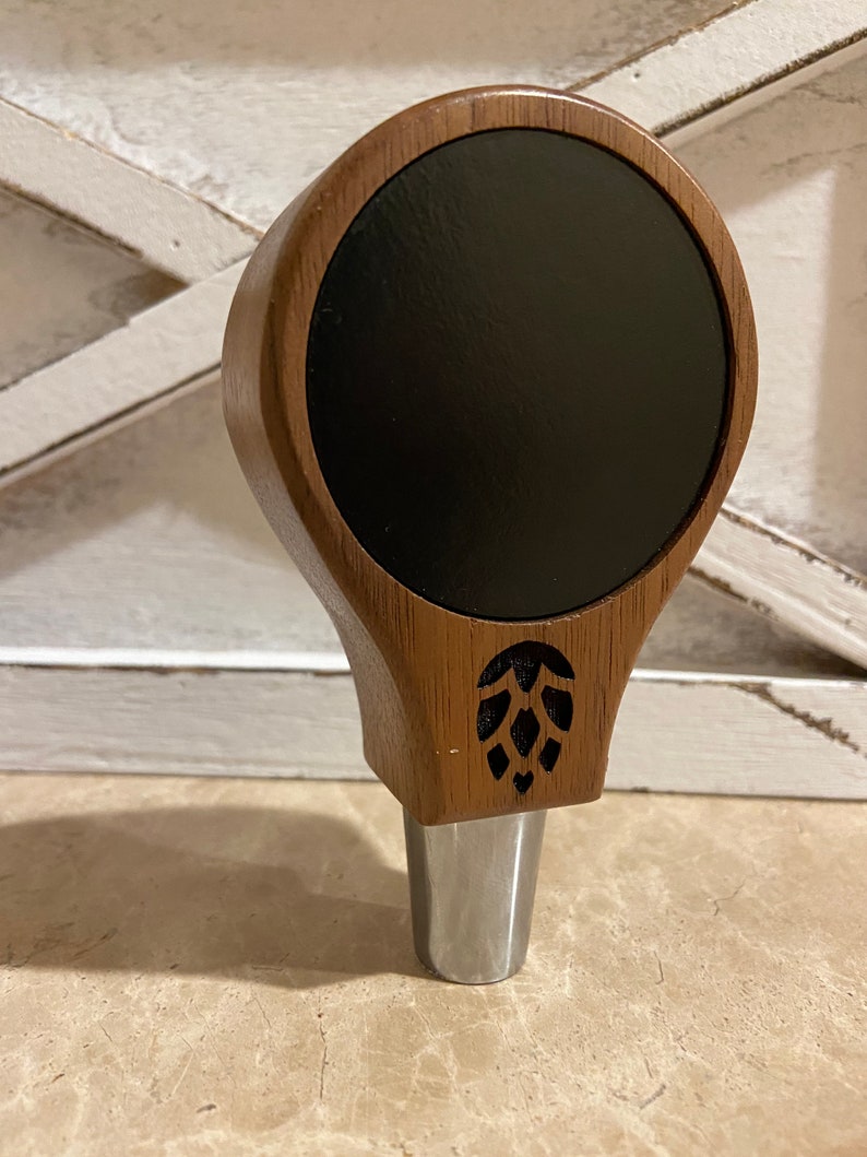 May include: A wooden beer tap handle with a black circular insert and a carved hop design. The handle is brown and has a silver metal base.