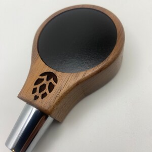 May include: A wooden beer tap handle with a black circular top and a carved hop leaf design. The handle has a chrome metal spout.