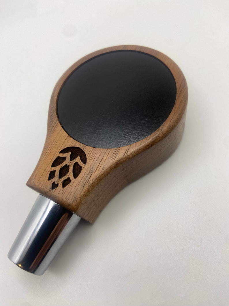May include: A wooden beer tap handle with a black circular top and a carved hop leaf design. The handle has a chrome metal spout.