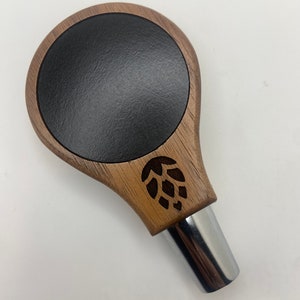 May include: A wooden beer tap handle with a black circular insert and a carved hop design. The handle is made of dark wood and has a chrome metal base.