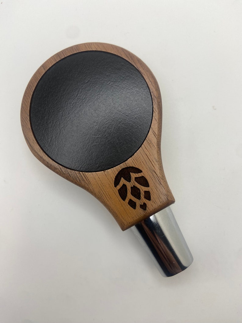 May include: A wooden beer tap handle with a black circular insert and a carved hop design. The handle is made of dark wood and has a chrome metal base.