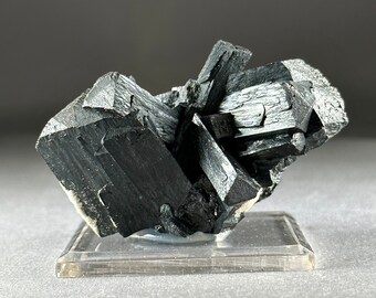 May include: A cluster of black, metallic, crystal formations on a clear acrylic display stand. The crystals are sharp and angular, with a glossy finish.