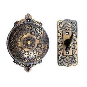 May include: Antique brass doorbell with an ornate design. The doorbell has a round face with a circular button in the center. The button is surrounded by a decorative pattern. The doorbell is attached to a rectangular brass plate with a similar ornate design.