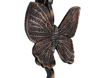 Puede incluir: Un llamador de puerta de mariposa de bronce con detalles intrincados. La mariposa tiene un esquema de color negro y marrón y está en posición de vuelo. El llamador está unido a un gancho de metal.