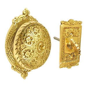 May include: A gold-toned doorbell with an ornate design. The doorbell has a round, embossed face with a floral pattern and a small, round button in the centre. The doorbell is attached to a rectangular, ornate plate with a lever.