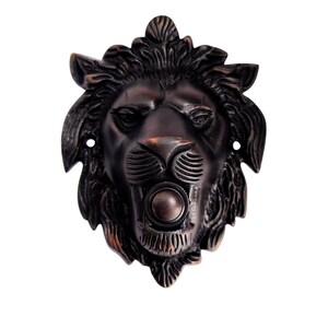 Può includere: Un copri campanello in metallo color bronzo scuro a forma di testa di leone con criniera e tratti del viso dettagliati. Il leone presenta un pulsante circolare situato al centro della bocca e due piccoli fori di montaggio sui lati.