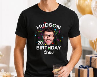 May include: A black short-sleeved t-shirt featuring a graphic print with the text "HUDSON 20th BIRTHDAY Crew" in white letters. The design includes a central photographic portrait of a smiling person wearing glasses, surrounded by colorful confetti.