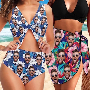 May include: Two women wearing swimwear with a tropical print featuring a repeated pattern of a man's face. The swimwear is blue, white, and pink with green leaves. The woman on the left is wearing a bikini top and high-waisted bikini bottoms. The woman on the right is wearing a black bikini top and a sarong with the same tropical print as the bikini.