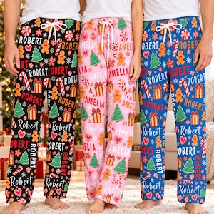 May include: Three pairs of pajama pants featuring a repeating Christmas pattern of gingerbread people, candy canes, gift boxes, and pine trees. The black pants feature the name Robert, the pink pants feature Amelia, and the blue pants feature Robert.