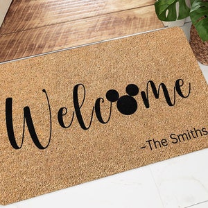 May include: A brown coir doormat with the text "Welcome" in black cursive lettering. A black Mickey Mouse silhouette is incorporated into the word "Welcome". The text "-The Smiths-" is printed in black at the bottom right corner of the doormat.