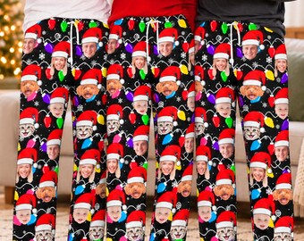 May include: Black pajama pants featuring a repeating pattern of colorful Christmas light bulbs and faces of people and pets wearing Santa hats. The pants have a white drawstring waist and are decorated with white snowflakes throughout the design.