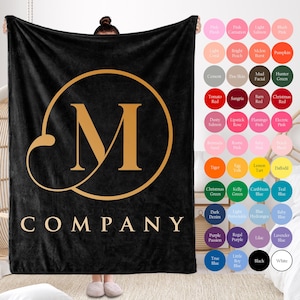 Peut inclure: Une couverture en polaire noire avec un cercle doré et la lettre "M" au centre. Le texte "COMPANY" est imprimé sous le cercle.