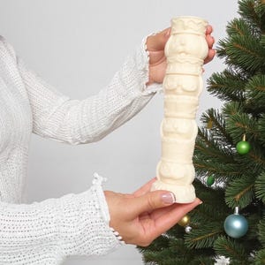 Könnte beinhalten: Ein cremefarbener, dekorativer Kerzenhalter mit gestapeltem Design, gehalten in der Nähe eines Weihnachtsbaums mit Ornamenten. Der Kerzenhalter weist eine Reihe detaillierter, geformter Figuren auf. Die Gesamtästhetik deutet auf einen festlichen, weihnachtlichen Artikel hin.
