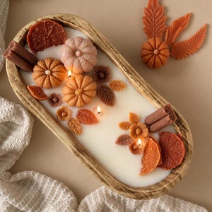 Peut inclure: Une bougie dans un bol en bois ovale contenant de la cire blanche et trois mèches allumées. La surface est décorée d'ornements en cire incluant des citrouilles orange et marron, des bâtons de cannelle, des tranches d'orange et des feuilles.
