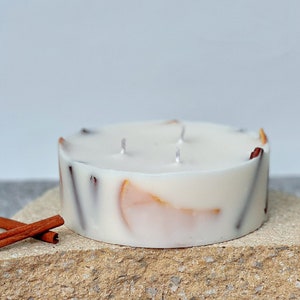 May include: A white candle with cinnamon sticks and orange peel embedded in the wax. The candle is sitting on a light brown stone surface.