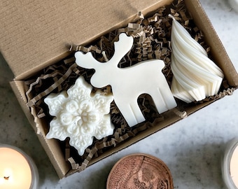 May include: A gift box with white holiday-themed candles and decorative soaps. The box contains a snowflake, a reindeer, and a Christmas tree-shaped soap. Lit candles and pinecones surround the box.