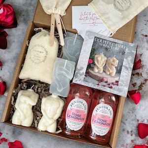 May include: A gift box filled with a variety of items, including two candles shaped like human figures, two bottles of rose lemonade, and two small cloth bags. The box is surrounded by red rose petals.