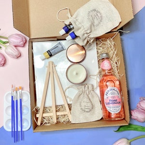 May include: A gift box filled with art supplies, including a small easel, paintbrushes, paint tubes, and a canvas. The box also includes a candle, a bottle of rose lemonade, and a small pouch.