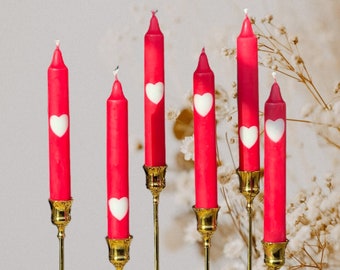 May include: Seven red candles with white heart designs are displayed on gold candle holders. The candles are arranged in a row on a light beige surface.