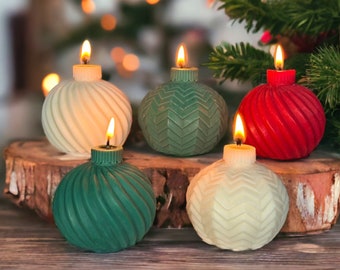 May include: Five Christmas ornament-shaped candles with lit wicks. The candles are in shades of white, green, and red, with textured surfaces. They are arranged on a wooden surface in front of a Christmas tree.
