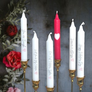 Könnte beinhalten: Fünf Kerzenständer mit weißen Kerzen und goldenen Kerzenhaltern. Die Kerzen haben verschiedene Botschaften darauf geschrieben, darunter "Happy Valentine's Day", "Forever & Always", "You are Enough", "Our First Valentine's Together" und "Will You Be My Valentine". Eine Kerze ist rot mit einem Herz-Ausschnitt.
