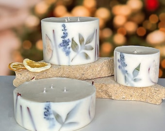May include: Three white candles with dried flowers and leaves embedded in the wax. The candles are different sizes and are arranged on a light brown surface.