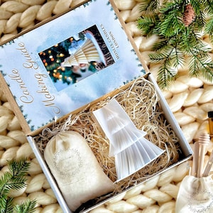 May include: A white box with a brown lid containing a candle making kit. The box is open and shows a white candle mould in the shape of a Christmas tree, a bag of wax flakes, and a small bag of wicks. The box is sitting on a white knitted blanket with a green pine branch in the foreground.
