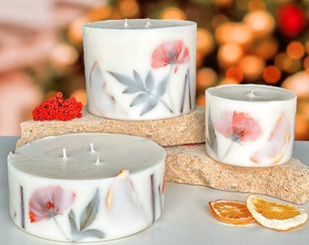 May include: Three white pillar candles with floral designs and dried orange slices. The candles are arranged on a stone surface with red berries in the background.