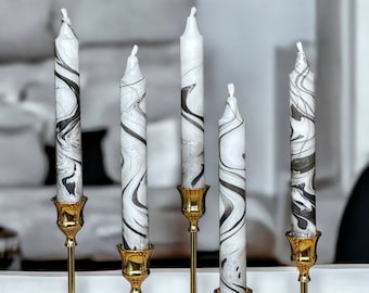 May include: Five white marble-patterned candles with black swirls, displayed on gold-colored candle holders. The candles vary in height, creating a visually appealing arrangement. The background is a blurred, neutral-toned interior.