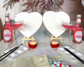 May include: A Valentine's Day paint and sip kit with two white heart-shaped canvases, two glasses of pink lemonade, two red candles, paintbrushes, paint, and two cards that say "Happy Valentine's Day!"