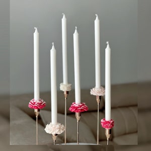 May include: Six white candles with pink and white flower candle holders. The candles are arranged in a row on a table.