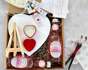 May include: A gift box filled with items for a creative art project. The box contains a heart-shaped canvas, a small easel, paintbrushes, paint, a candle, and two bottles of rose lemonade. The box is lined with brown paper shreds.