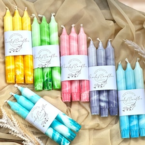 Könnte beinhalten: Eine Auswahl handgetauchter Kerzen in verschiedenen Farben, darunter Gelb, Grün, Rosa, Lila und Blau. Jedes Set ist mit einem weißen Etikett mit der Aufschrift "Hand Dipped in London" und "Henley Candles" versehen.