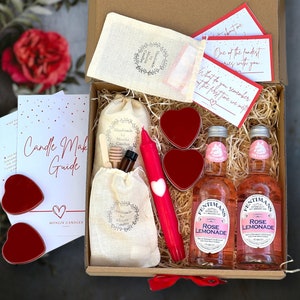 May include: A gift box filled with items for making candles, including a candle making guide, a bag of wax flakes, a jar of honey, a bottle of essential oil, a red candle, and two small red heart-shaped candles. The box also includes two bottles of rose lemonade.