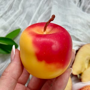 May include: A hand holds a red and yellow apple with a brown stem. The apple's colours blend from yellow to red. Sliced apples are visible in the background.