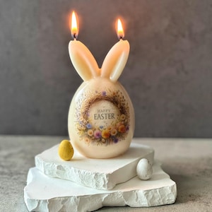 Könnte beinhalten: Eine weiße, kaninchenförmige Kerze mit einem floralen Kranzdesign und dem Text "Frohe Ostern". Die Kerze hat zwei brennende Dochte und steht auf einem weißen Steinsockel.