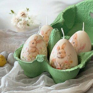 Może przedstawiać: Cztery białe, jajowate świece z ilustracjami królików w zielonym kartonie na jajka. Każda świeca przedstawia inny wzór królika z kwiatowymi akcentami.