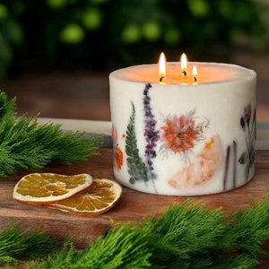 May include: A white candle with dried flowers embedded in the wax. The candle is lit and has three wicks. The candle is sitting on a wooden surface with dried orange slices.