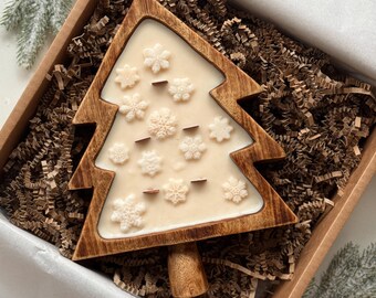 May include: A Christmas tree-shaped candle with snowflake accents, set in a wooden frame. The candle is cream-colored and rests in a cardboard box with shredded paper. The tree has a wooden trunk.